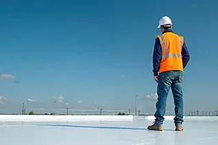 Roofer in orange vest, protective hard hat stands on roof coated with reflective paint. Man inspects flat commercial roof for energy efficiency, renovation. Contractor looks away at clear blue sky.