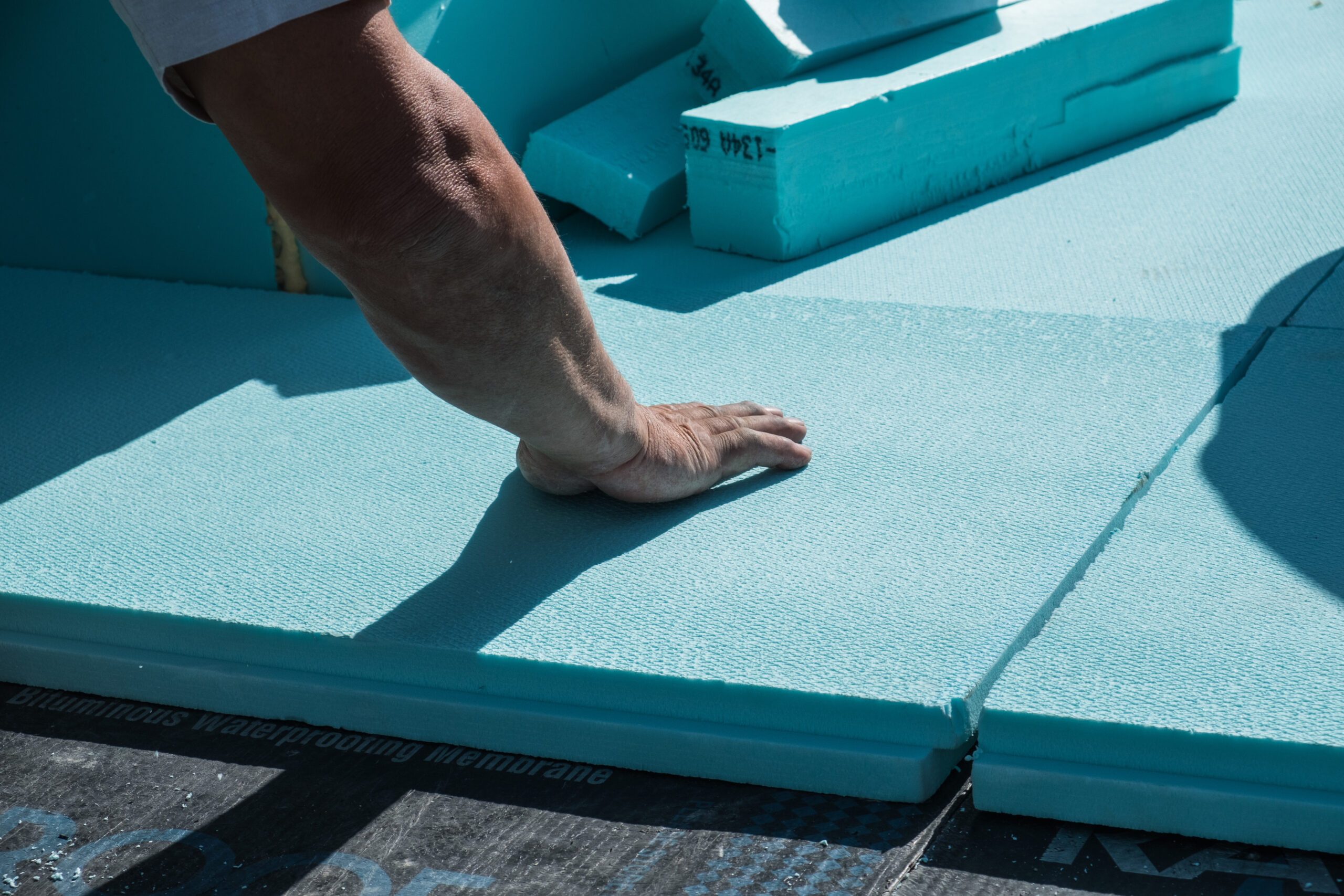 A roofer installs blue XPS polystyrene insulation boards on a flat roof