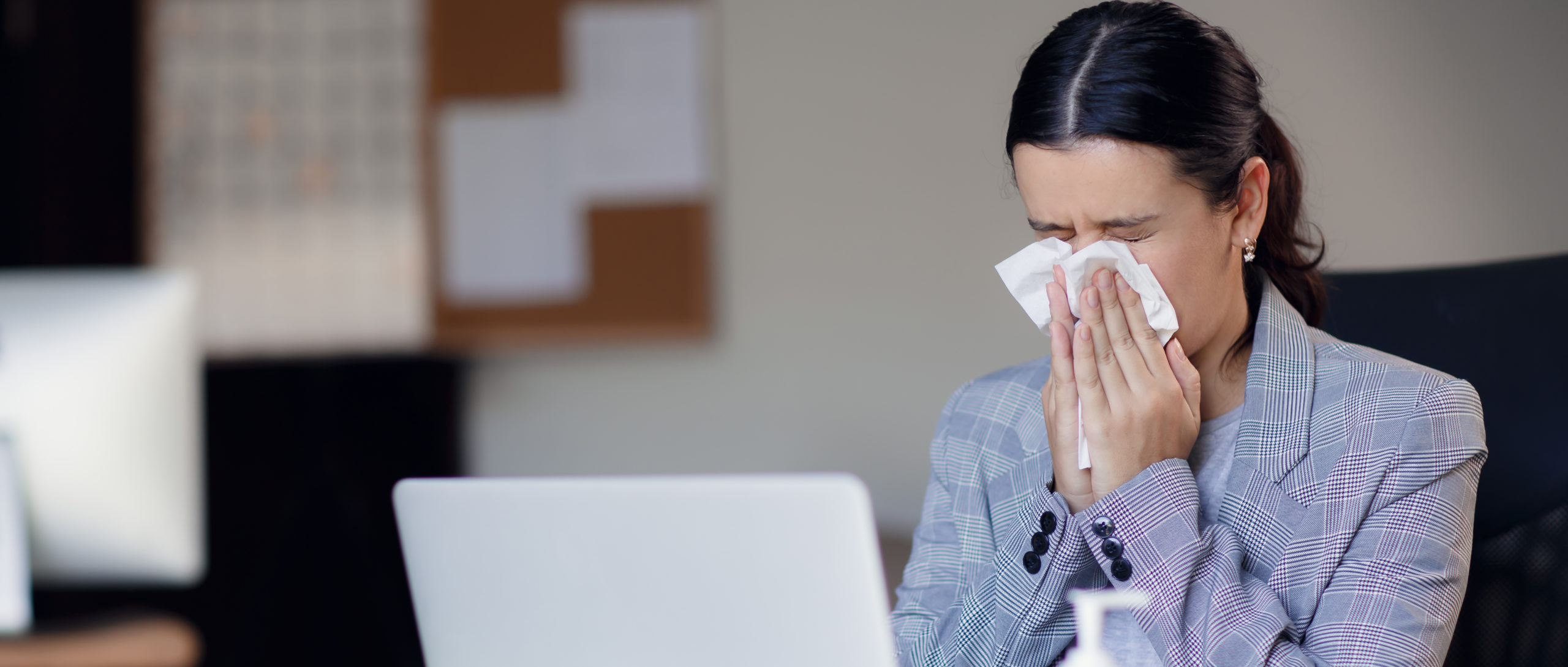 photo of a sick female employee sneezing into a tissue while working on a laptop