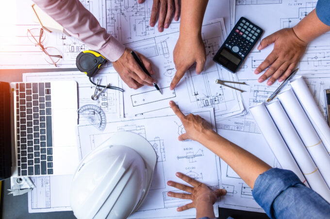 three engineers are pointing at a specific spot of a blueprint while another engineer attempts to calculate costs based on the blueprint