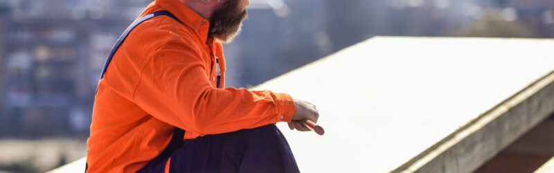a roofer wears orange protective gear and holds a tool as he looks at a white flat roof