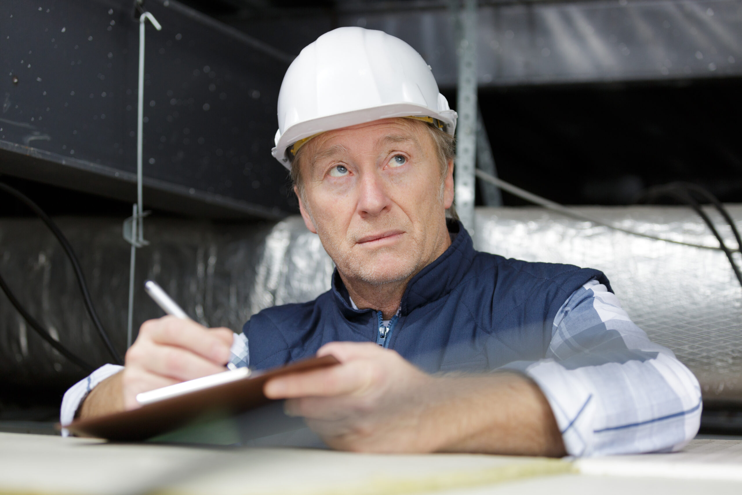 A white/Caucasian man wearing a white hard hat holds a clipboard and a pen during a roof inspection