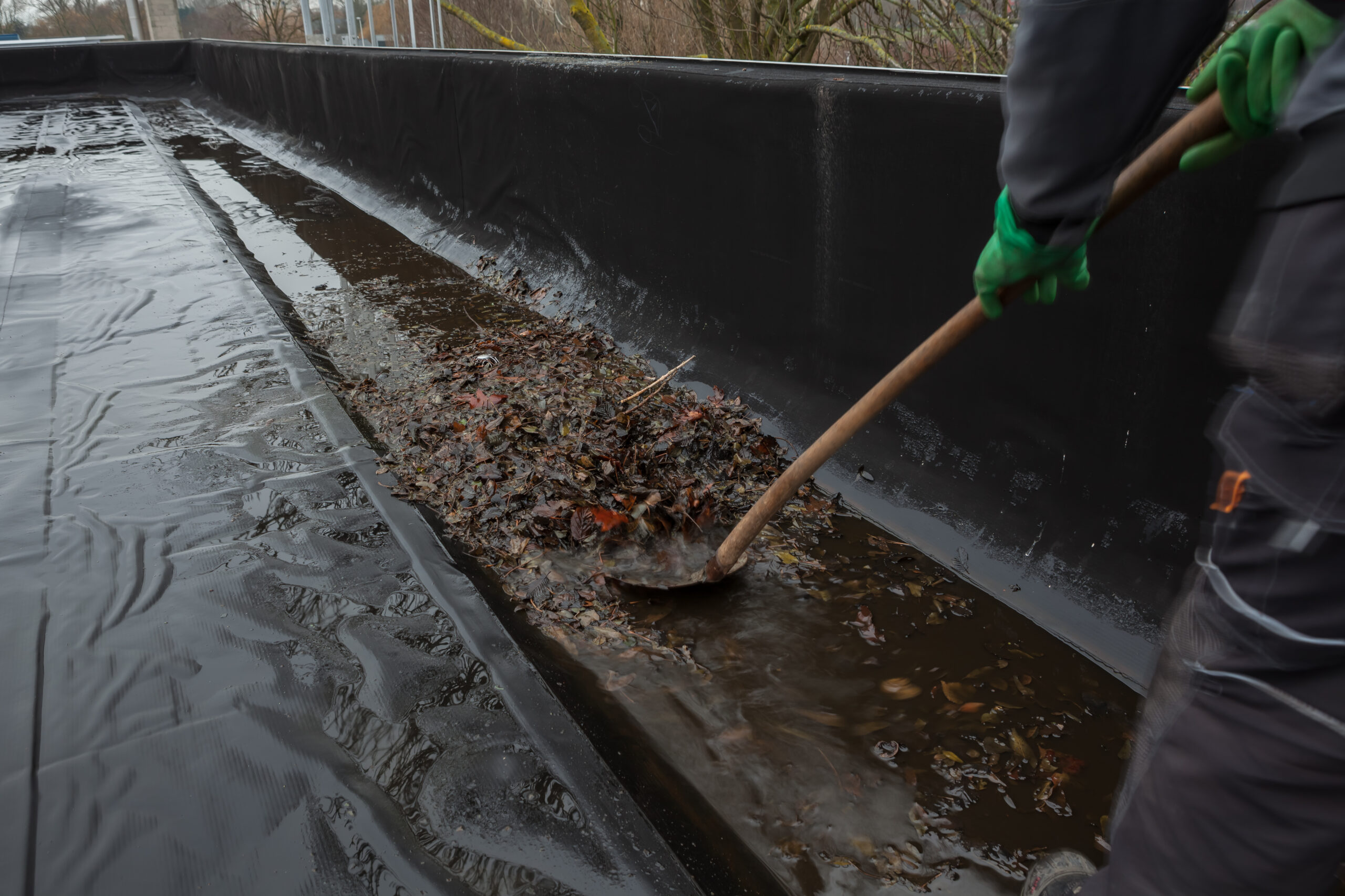 A man wearing protective clothing uses a shovel to remove pooled water and accumulated leaves from a black flat PVC roof