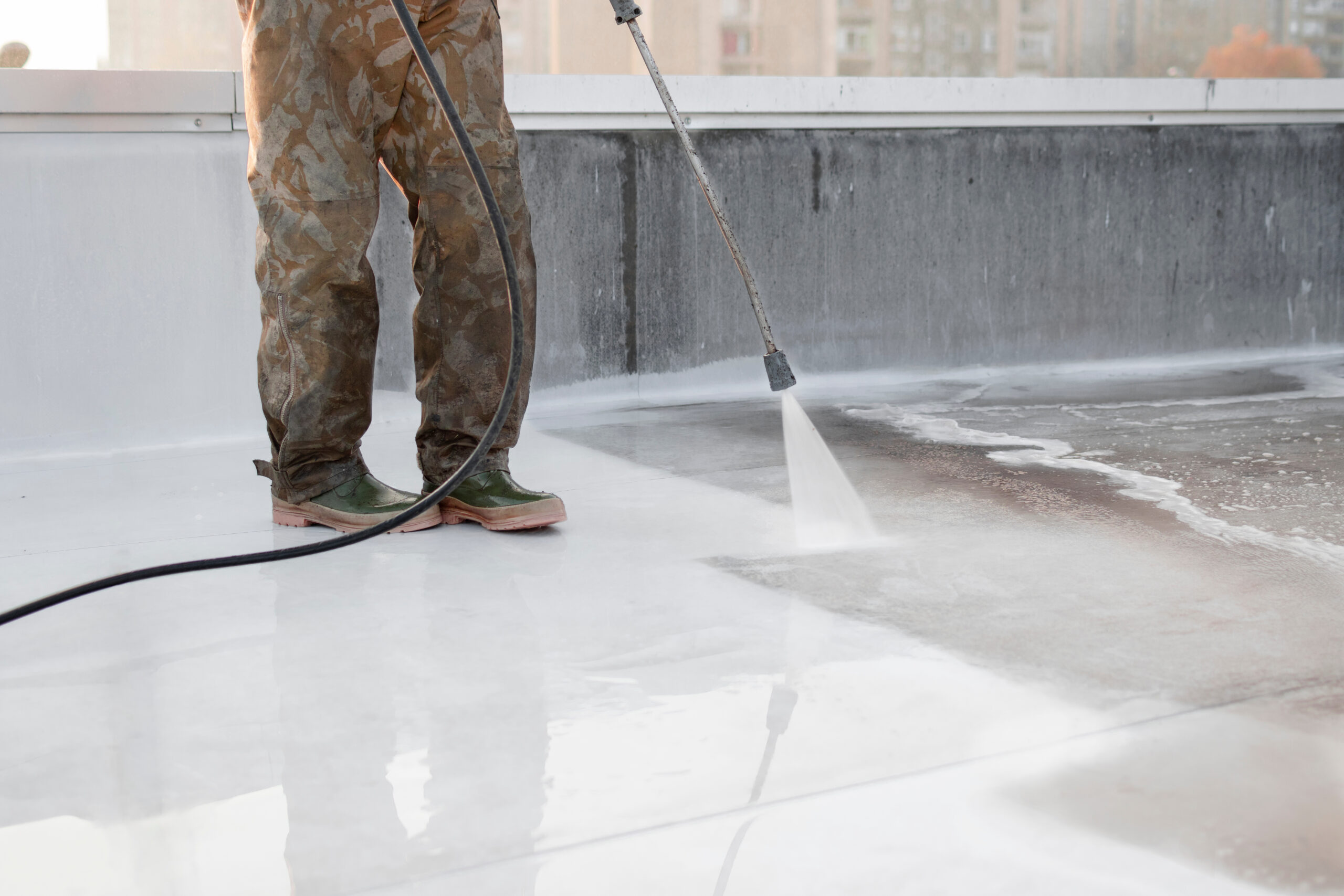A man wearing camoflauge pants is using a pressure washer on a low setting to clean a white flat roof